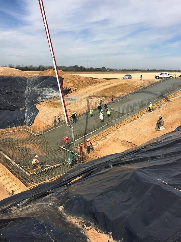 Construction workers installing black liner in a large excavation beside a roadway under a cloudy sky