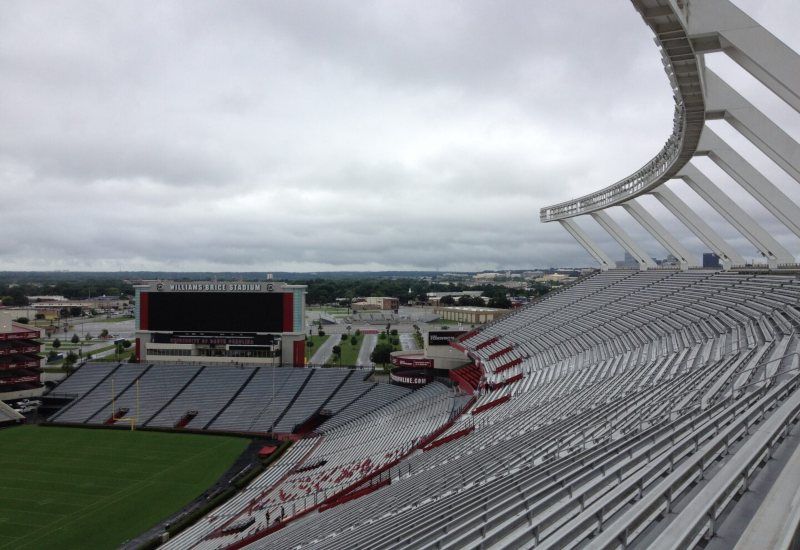 Empty stadium stands under a cloudy sky, with a large scoreboard and green field.