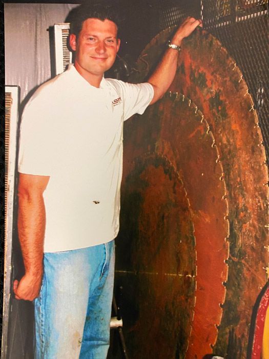 Man standing beside a large circular wooden disk, resting one hand on it indoors.