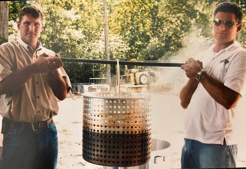 Two men holding a suspended metal basket over a steaming outdoor fire pit
