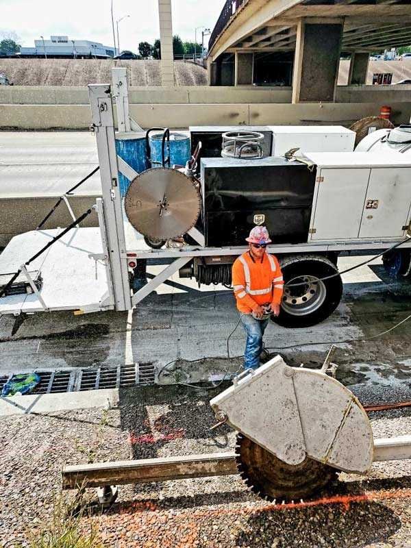 Worker in orange safety vest beside a utility truck and saw blade on a construction site