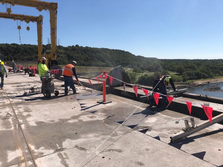 Workers in safety vests at a construction site with red caution tape beside a waterway