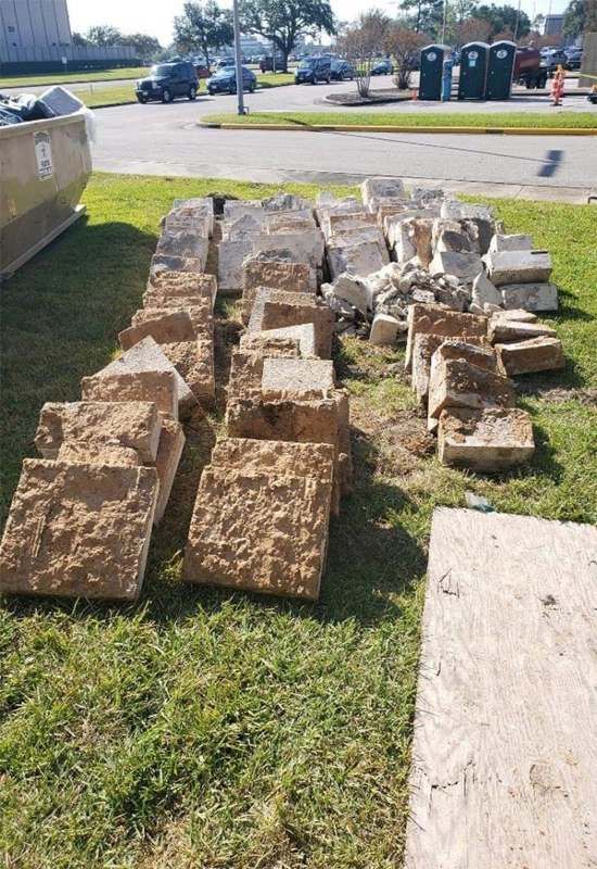 Stacks of tan stone or concrete blocks on a grassy curb beside a sidewalk and street, with portable toilets in the background
