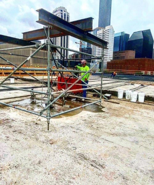 Worker in neon safety vest standing on rooftop scaffolding beside a large beam, city skyline behind.