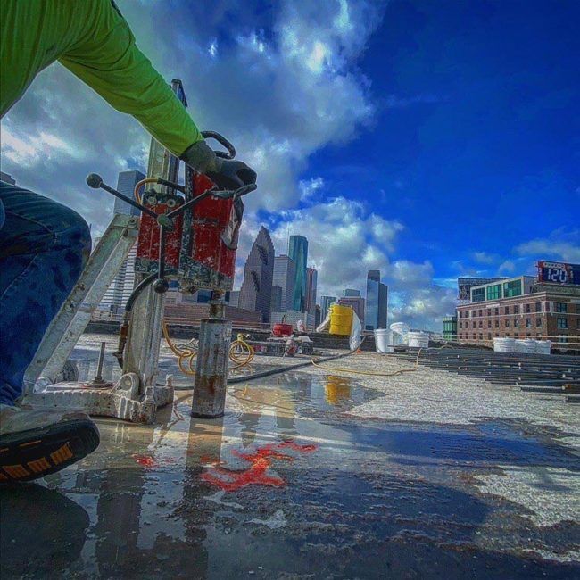 Worker on a rooftop with city skyline and blue sky, using a hose near puddles and construction equipment.