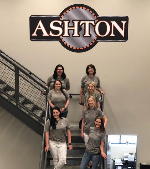 Six people on an indoor staircase beneath an ASHTON sign, posing and smiling.