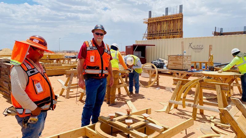 Construction workers in orange safety vests and hard hats at a sunny building site with wooden framing.