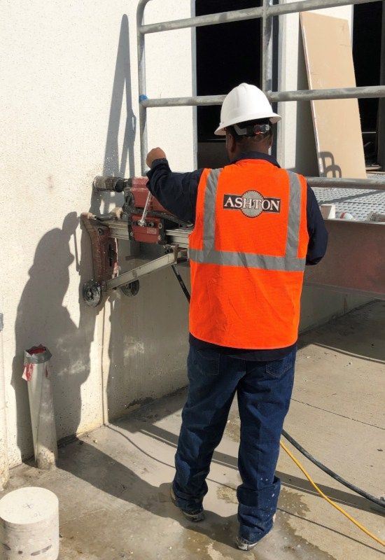 Construction worker in orange vest and hard hat using a wall-cutting saw on a concrete wall.