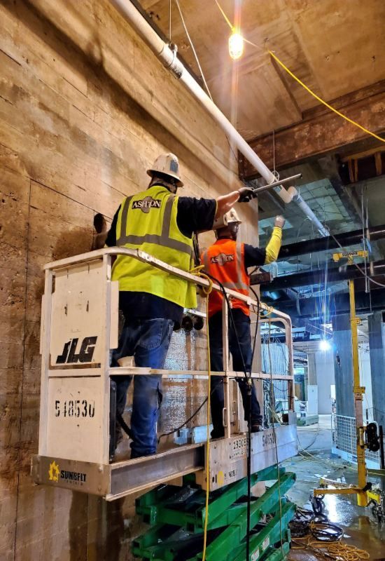 Two construction workers on a lift installing ceiling pipes in a dim indoor site.