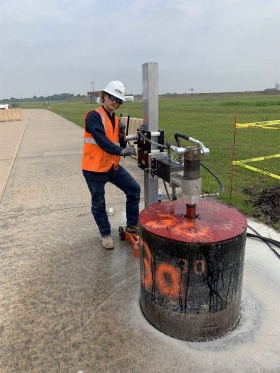 Worker in safety gear operating a device over a smoking burn barrel beside a roadway.