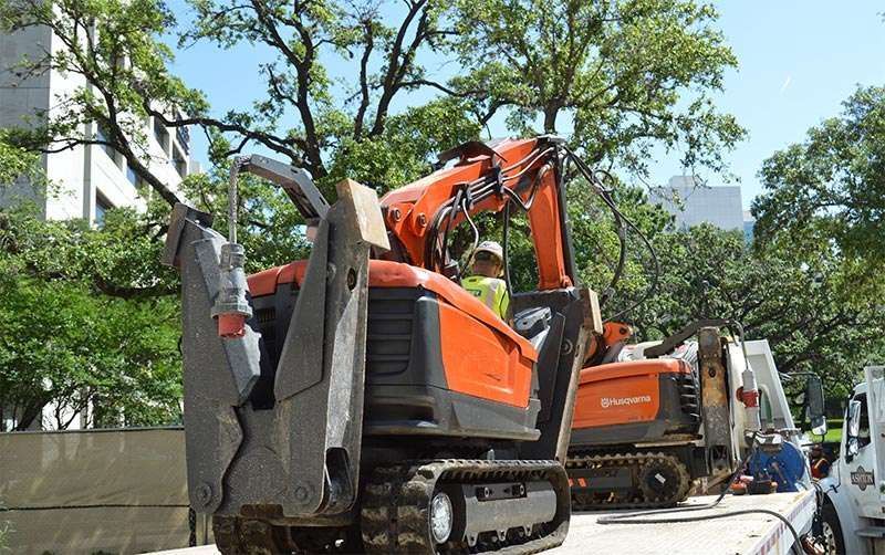 Orange excavator beside a concrete wall, lifting debris in a tree-lined urban area