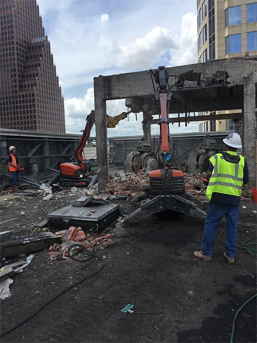 Construction workers using excavators to clear debris from a burned rooftop parking structure under cloudy skies