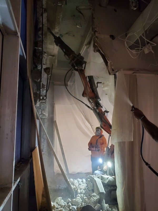 Collapsed hallway with rubble; a worker in an orange vest inspects damage with a flashlight.