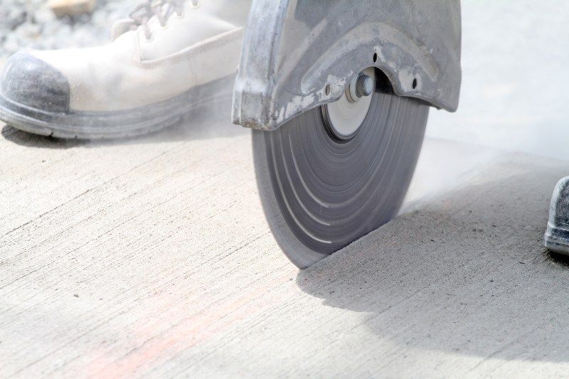 Concrete saw cutting a pavement slab, with a worker’s boot visible nearby.