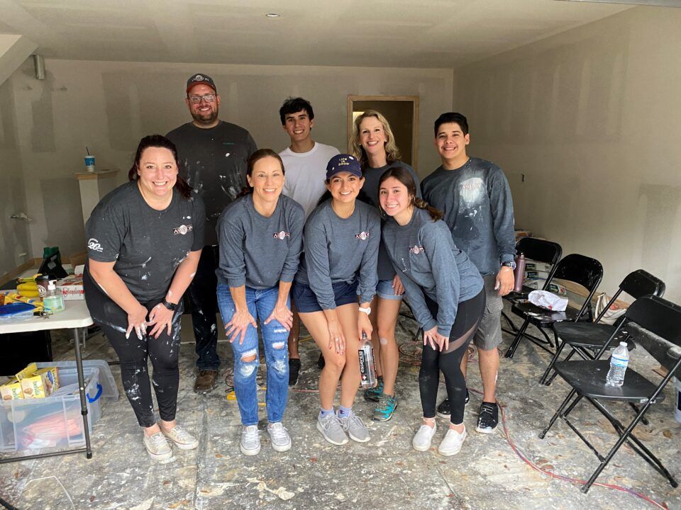 Seven people pose together in a bare room with unfinished drywall and tools on the floor.