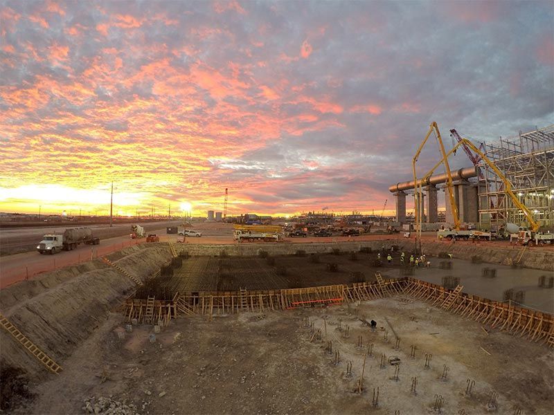 Construction site at sunset with cranes, excavator, and a large excavation pit under a pink sky