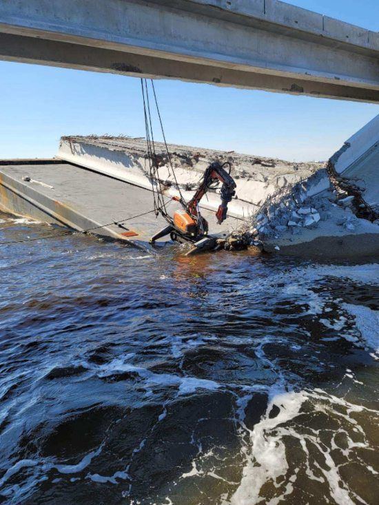 Person dangling beneath a collapsed bridge over choppy water, with debris and cables around them