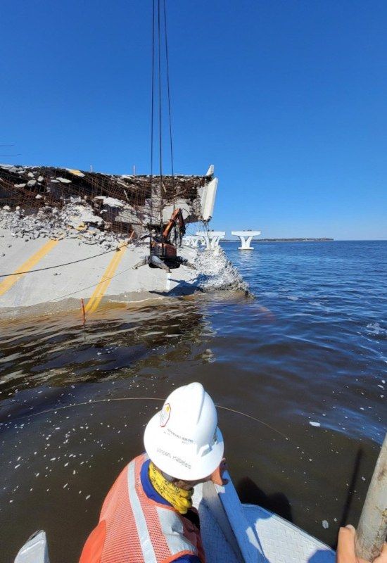 Person in a white hard hat riding a boat toward a damaged rocky shoreline under a clear blue sky