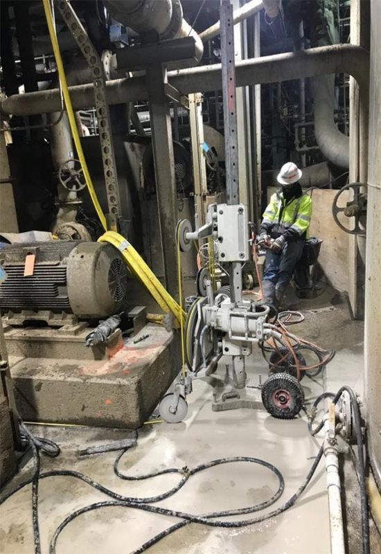 Industrial worker in a hard hat inspecting machinery in a dim factory area with cables and equipment.