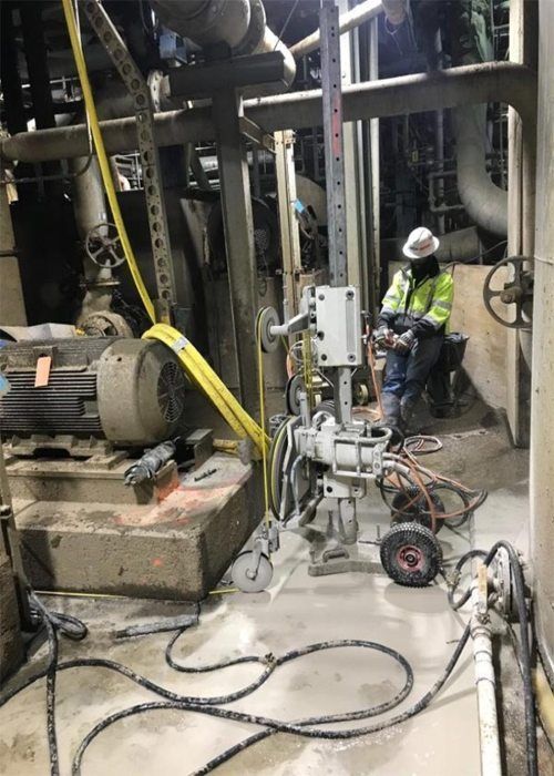 Worker in a hard hat seated near industrial machinery in a dim factory setting with cables on the floor