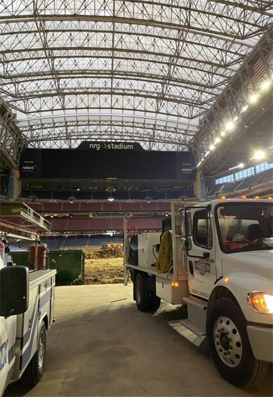 Trucks inside a stadium under a high arched roof, with dirt and debris in the field below.
