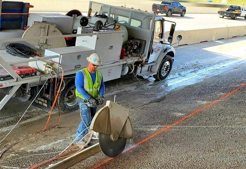Worker in safety vest operating a road saw beside a street repair truck on a covered roadway.