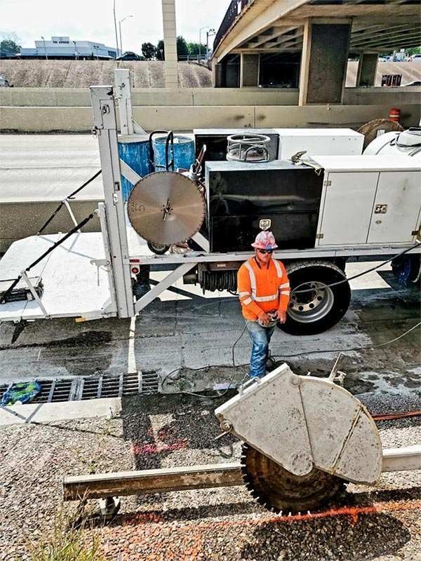 Construction worker in orange vest beside a road machine under an overpass, with traffic barriers and debris.
