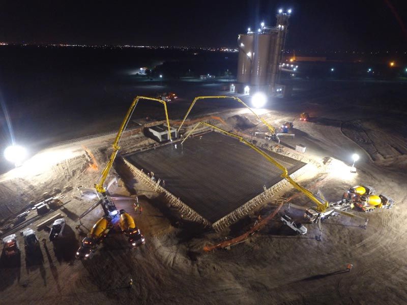Night construction site with cranes lifting a large concrete slab under bright floodlights