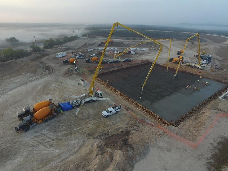 Aerial view of a construction site with concrete pumps, trucks, and a large foundation slab under construction.
