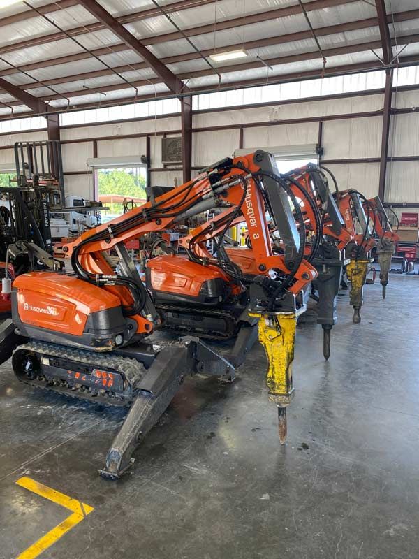 Row of orange excavators in a warehouse, with yellow attachments and black tracks on a concrete floor.