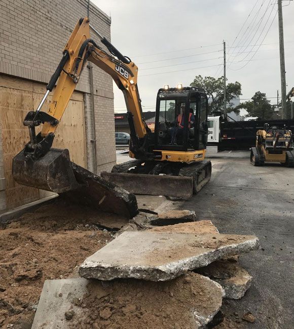 Yellow excavator breaking up concrete debris beside a building on a wet street