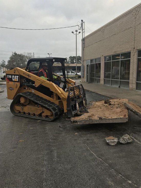 Yellow skid steer moves dirt near a beige building on a wet pavement.