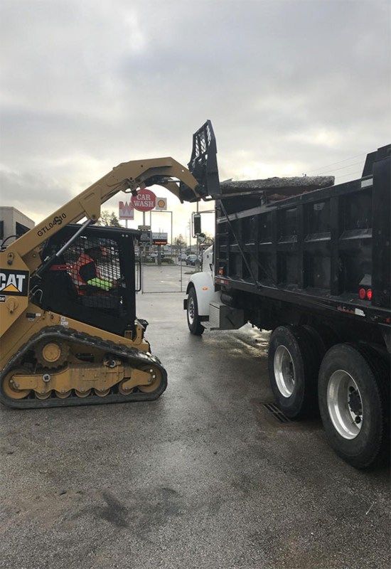 CAT skid steer loading debris into a black dump truck on a wet lot