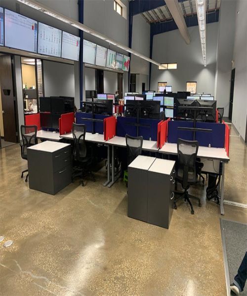 Open-plan office with rows of desks, monitors, red dividers, and black rolling chairs