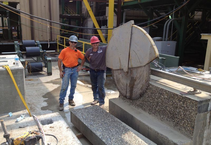 Two construction workers stand beside a large stone-cutting machine at an industrial site.