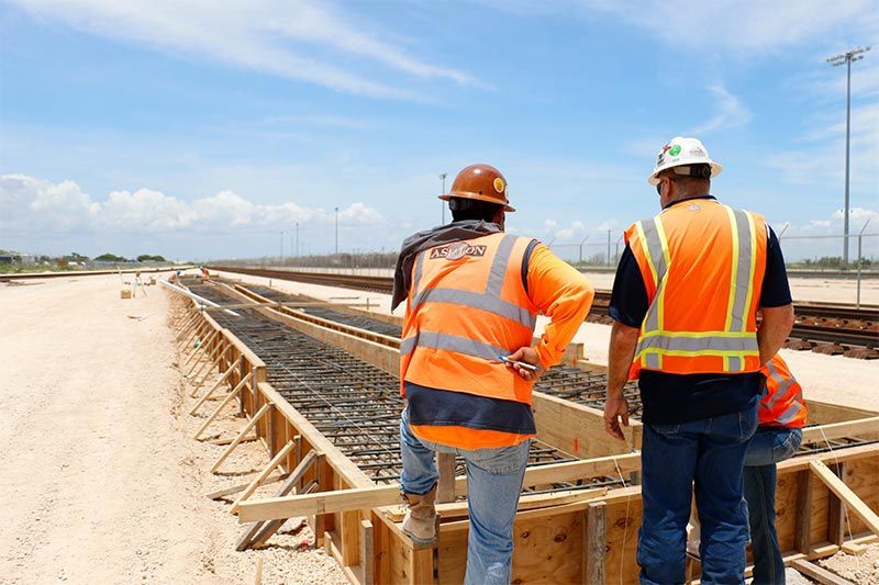 Two construction workers in safety vests and hard hats inspect a rebar foundation at a roadwork site