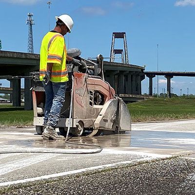 Worker using a concrete saw on a road near a bridge, cutting a white line outdoors.