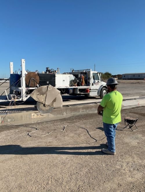 Worker in neon shirt stands by a utility truck and large reel in an open lot under a clear blue sky.