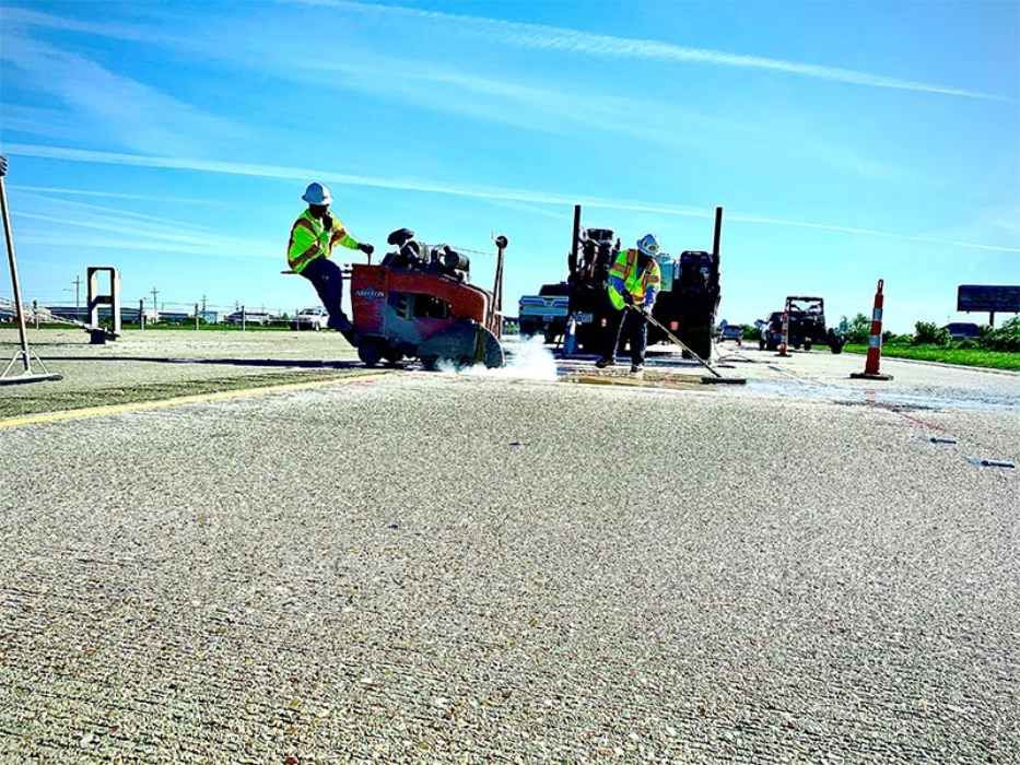 Workers in neon vests using a paving machine on a road under a bright blue sky