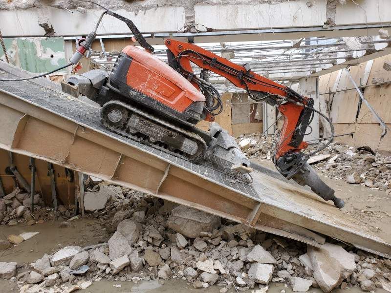 Orange excavator demolishing a collapsed concrete building, with rubble and debris around it