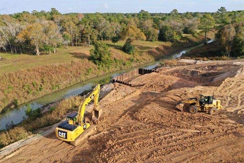 Excavators grading a large dirt site beside a canal, with trees in the background.