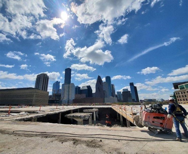 Construction site with city skyline under a blue sky; worker stands near an orange truck.