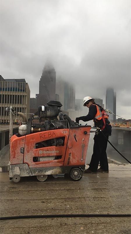 Worker standing beside orange construction machinery on a wet city street under a cloudy skyline