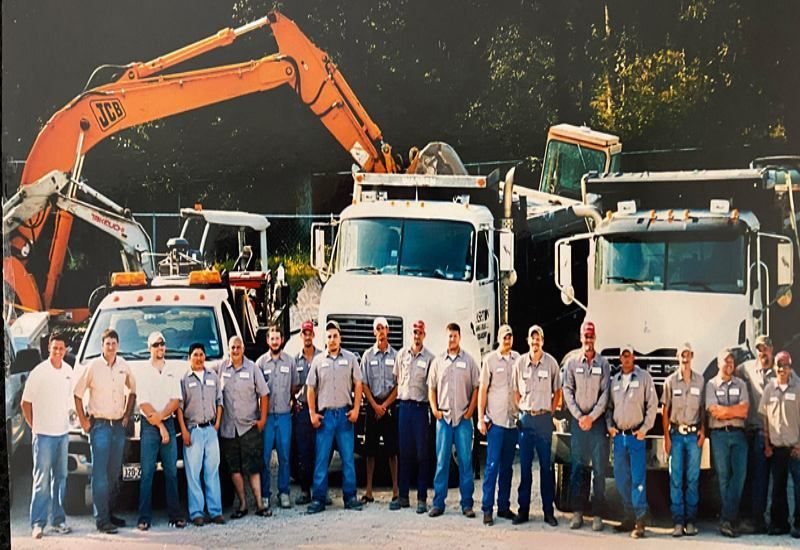 Construction crew posing in front of dump trucks and an excavator at a work site
