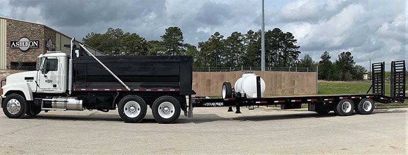 White dump truck towing a flatbed trailer with tanks in a parking lot under cloudy skies