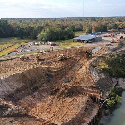 Aerial view of a large dirt excavation site with heavy machinery, a building, and a wooded area nearby.