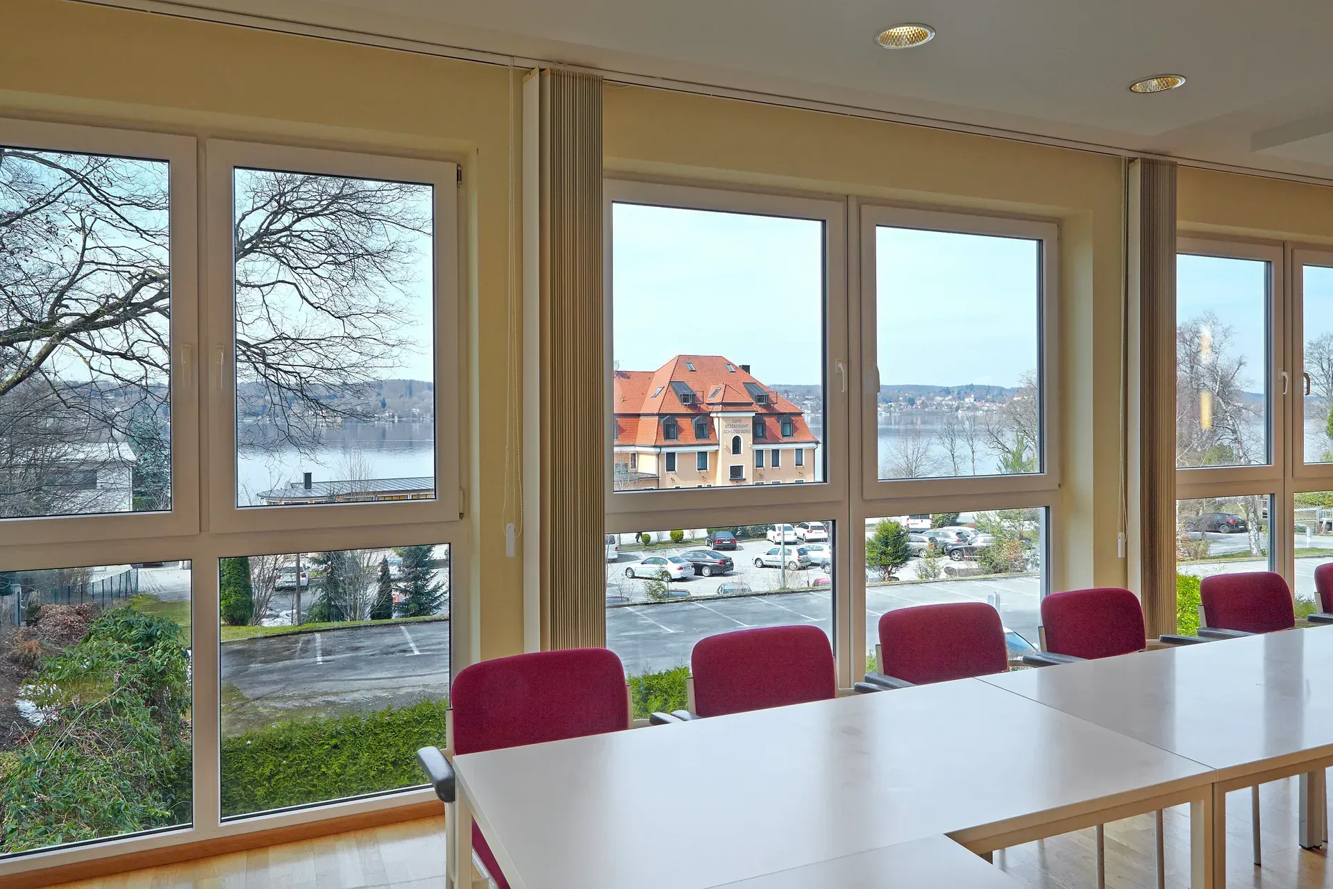 Conference room with large windows overlooking a lake and building; red chairs at a long table.