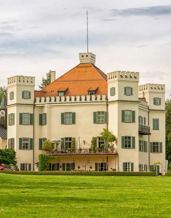 Historisches, weißes, schlossähnliches Gebäude mit orangefarbenem Dach neben einer grünen Rasenfläche und Bäumen