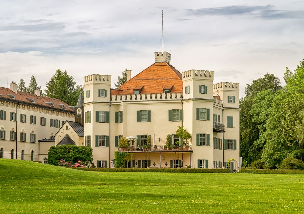 Historisches, weißes, schlossähnliches Gebäude mit orangefarbenem Dach neben einer grünen Rasenfläche und Bäumen