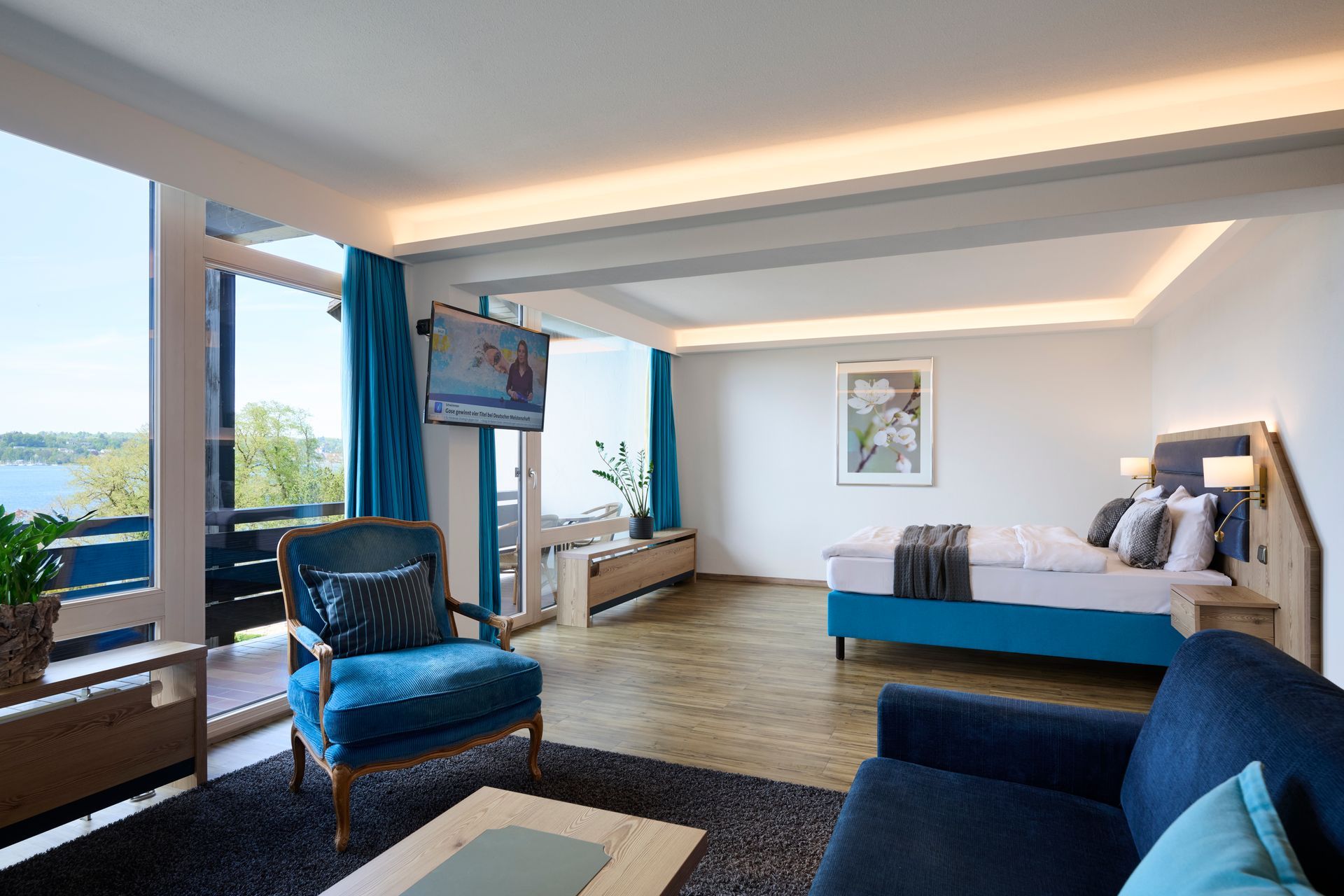Hotelzimmer mit blauem Sofa, Holztisch, Blumenstrauß und Seeblick vom Balkon.